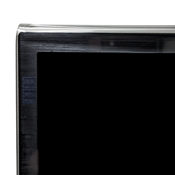 A close-up view shows the top corner of a flat-screen television. The screen is black, and the frame is also black with a metallic edge. The background is completely black, focusing attention on the corner of the TV. Screen Protector is attached with velcro tab at the front of the TV, behind the screen protector.
