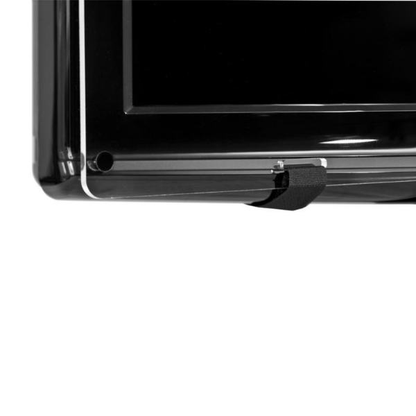 A close-up view from the side and slightly below shows the bottom corner of a clear acrylic TV screen protector attached to a black flat-screen television. A black Velcro strap wraps around the back of the TV and fastens to the protector. Part of the black TV screen and the side of the TV casing are visible. The background is plain white.