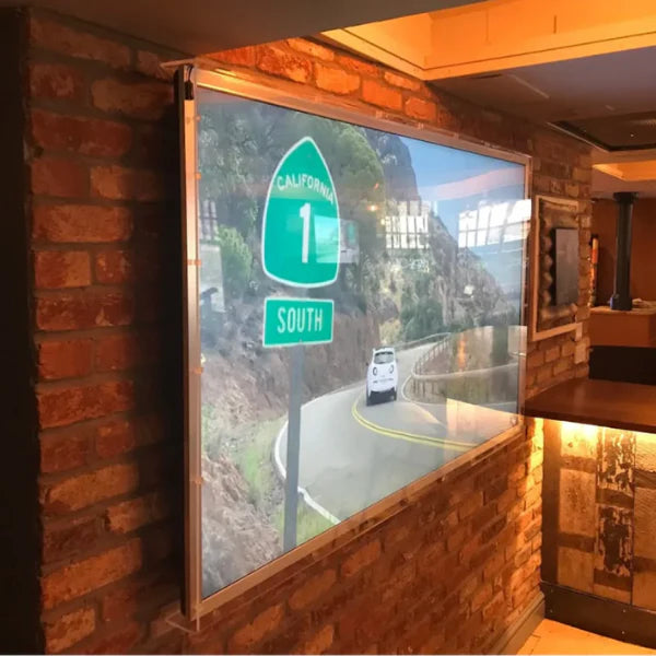 See the clarity and vibrant display through our TV screen protector. This image shows a large TV mounted on a brick wall, playing a vivid scene of a drive down California Highway 1 South, highlighting the undisturbed viewing experience.
